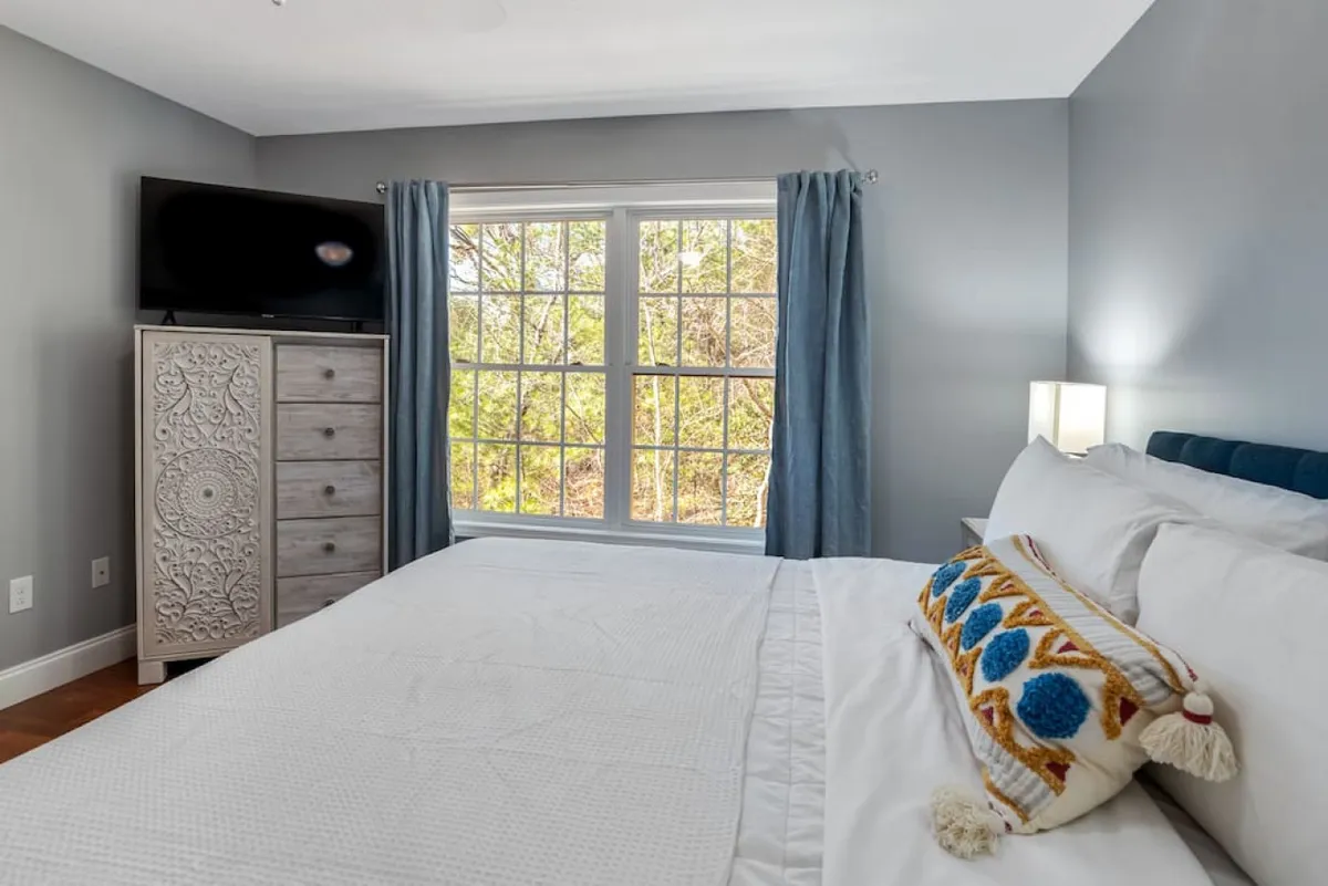 A serene bedroom scene with a bed adorned with a decorative pillow, a large window streaming in natural light, a dresser with a decorative design, and a television. The room feels cozy, bright, and perfectly suited for unwinding or enjoying views of nature.