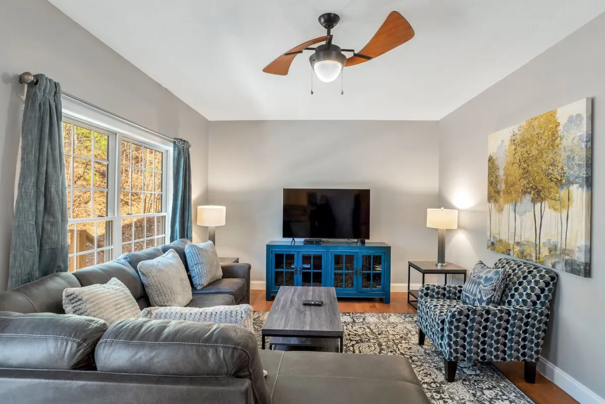 The image showcases a spacious, inviting living room with a plush sofa, a stylish blue cabinet, vibrant artwork, a warm lamp, and a large TV, creating a cozy and welcoming space perfect for relaxing or gathering with loved ones.