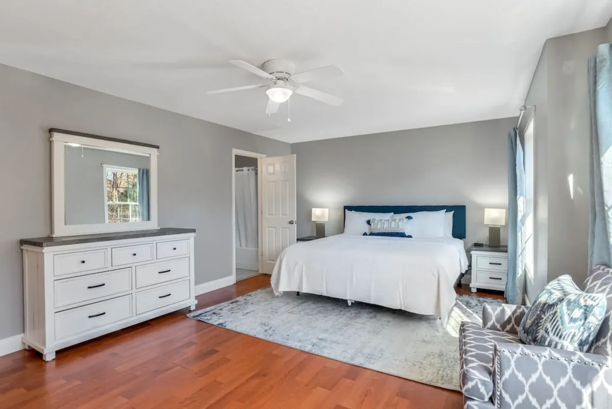 A soothing bedroom scene featuring a comfortable bed, a dresser, an armchair, and a window. The mood is serene, ideal for restful nights, cozy mornings, or quiet reading by natural light.