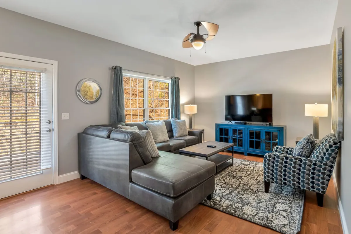 This image captures a cozy living room centered around a plush sofa, an armchair, coffee table, and large window that brings in natural light. The TV is positioned for easy viewing from the sofa, and the arrangement encourages relaxation, socializing, or enjoying a movie night.