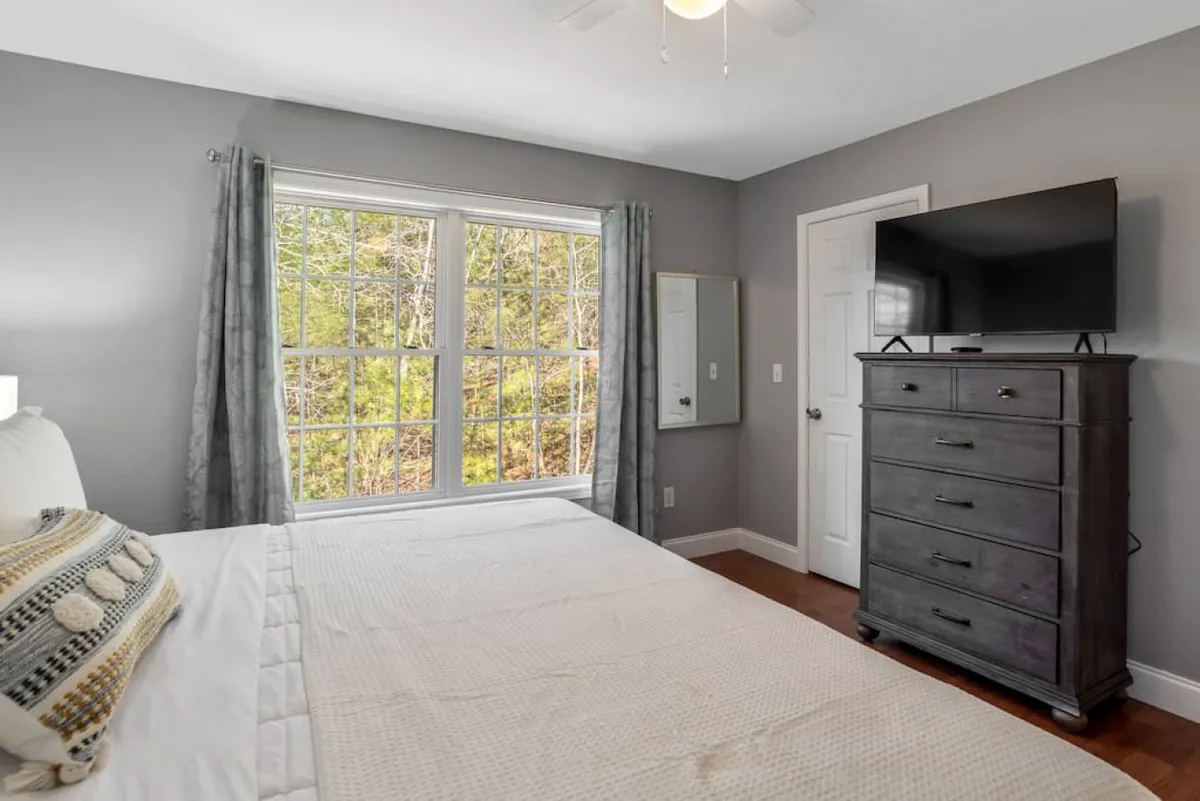 A serene bedroom featuring a comfortable bed positioned near large windows, allowing ample natural light to flood the room. The TV on the dresser offers additional comfort, and the overall mood is tranquil and inviting.