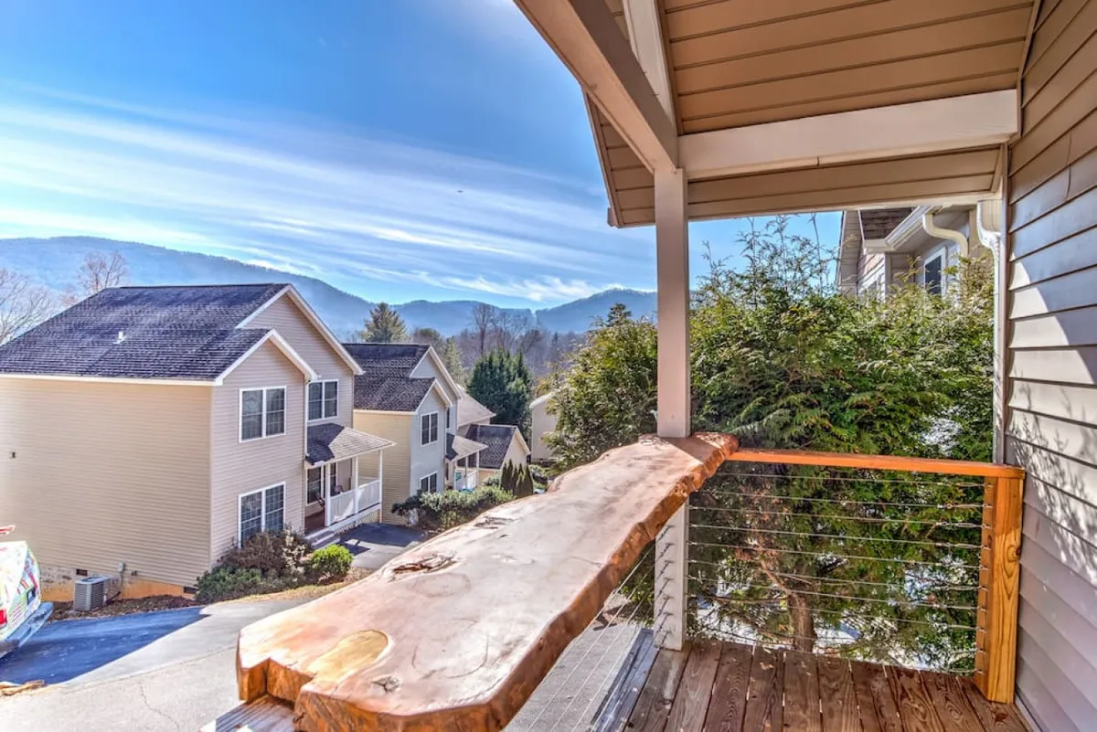 This image presents a tranquil mountain view from a vantage point with a wooden railing, overlooking neighboring houses. The vista includes sunrise or sunset lighting, evoking the peaceful, scenic environment guests return to after enjoying Asheville's urban offerings.