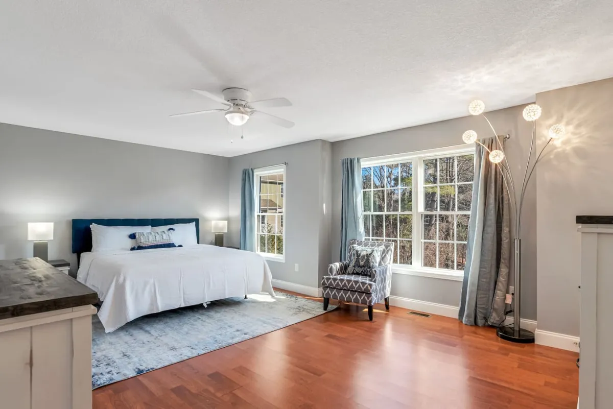 This image features a king-sized bed with white bedding, a modern accent chair, and a large window that allows abundant natural light to fill the space. The atmosphere is tranquil and inviting, suggesting a perfect spot for rest, reading, or enjoying the morning sunlight.
