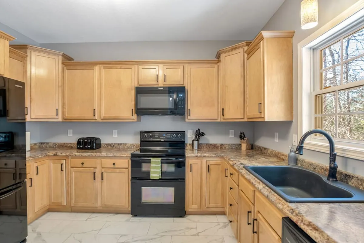 A warm, inviting kitchen scene featuring black appliances, granite countertops, and wooden cabinets. The kitchen window brings in extra light, and the setup is ideal for preparing snacks or connecting with others while cooking.