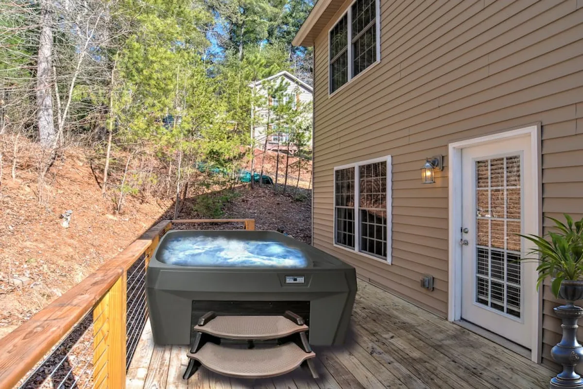 This image captures the exterior of the home, featuring a wooden deck with a hot tub in the foreground. The scene is tranquil and inviting, with sunset views providing a sense of relaxation after a day exploring downtown. The house's exterior wall and the surrounding natural setting are visible, suggesting a seamless transition between the adventures of Asheville and a peaceful home base.