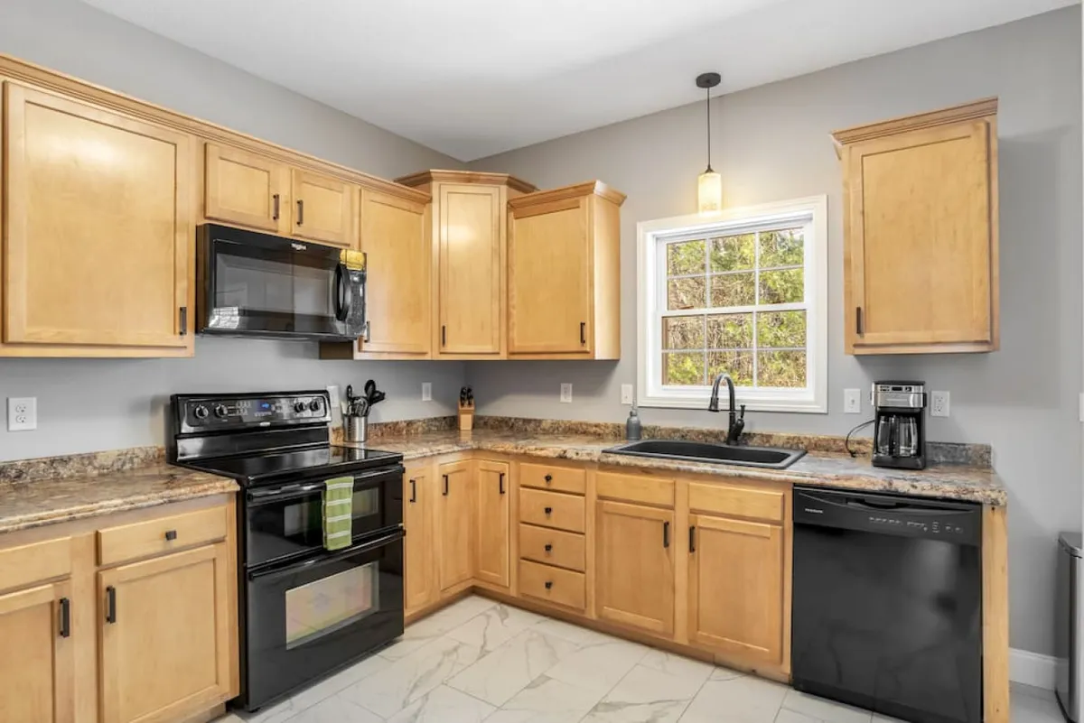 A welcoming kitchen scene with maple cabinets, black appliances, a granite countertop, and a window offering a view outside. The setup suggests a space for baking, cooking, or enjoying a morning coffee.